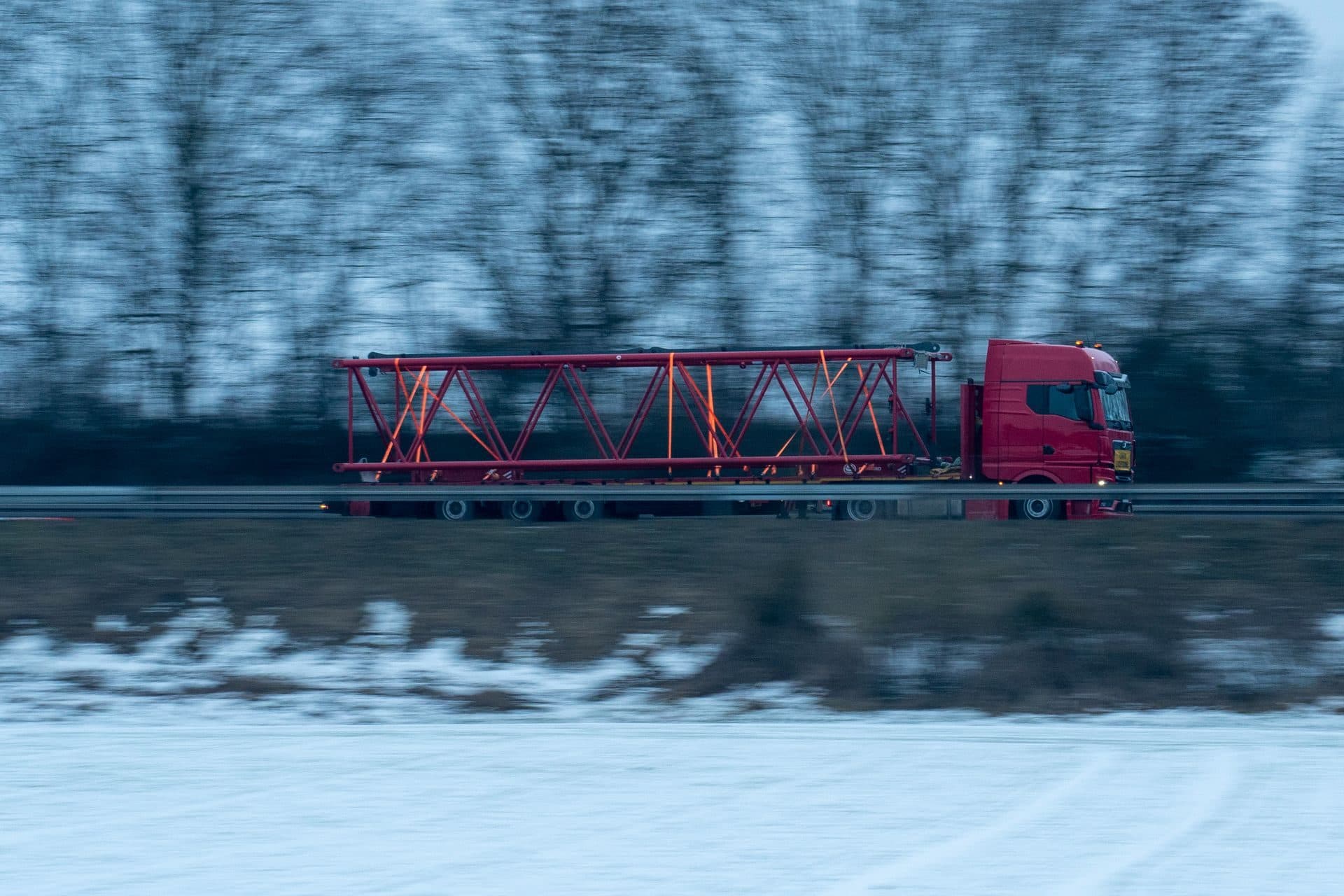 Red tractor hauling a red steel truss on a flatbed through snowy woods