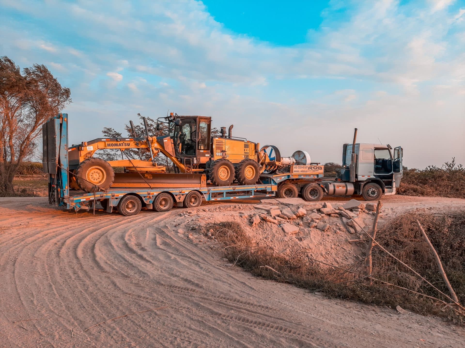 Lowboy trailer hauling a motor grader along a dirt road at dusk