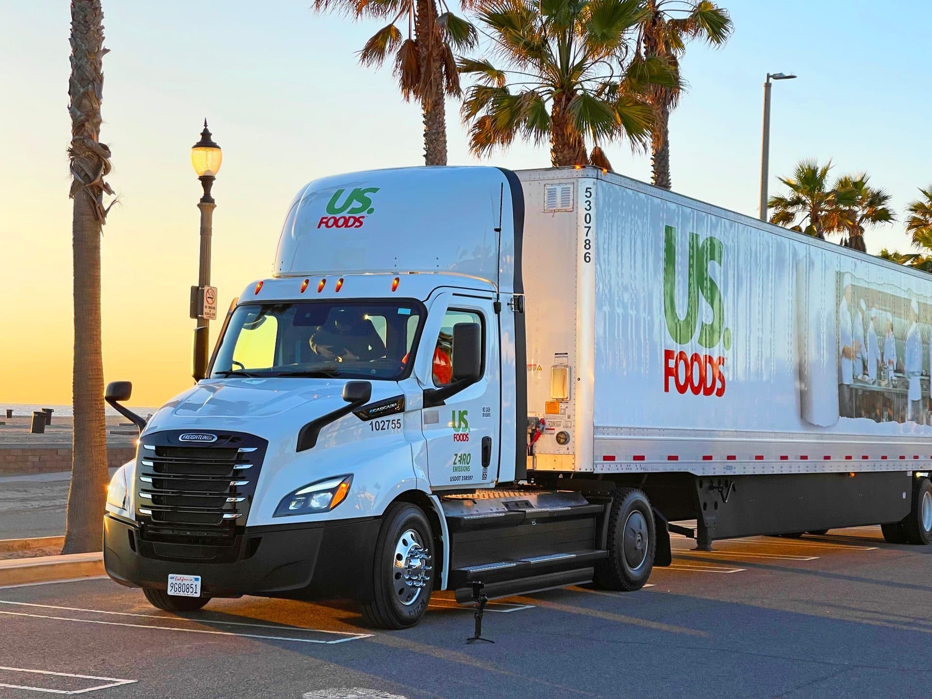 White tractor and US Foods refrigerated trailer parked beneath palm trees